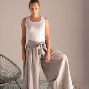 May include: A woman in a white vest top and light grey linen palazzo trousers with a matching tie belt. The wide-leg trousers are pleated. A green chair and a wicker basket with folded clothes are in the background.