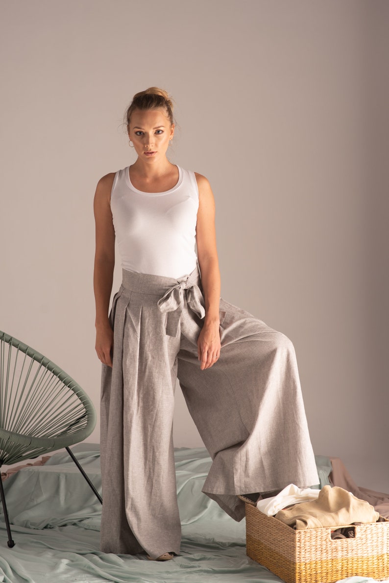 May include: A woman in a white vest top and light grey linen palazzo trousers with a matching tie belt. The wide-leg trousers are pleated. A green chair and a wicker basket with folded clothes are in the background.