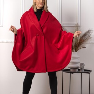 May include: A vibrant red cape coat with a double-breasted design. The coat features a wide collar and a flowing silhouette. The model is wearing black leggings and boots. The background is a white wall with decorative trim.