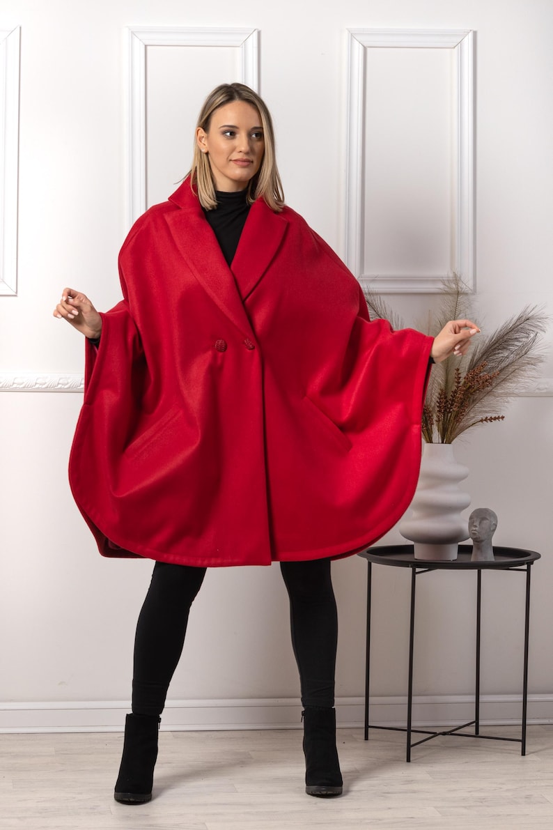 May include: A vibrant red cape coat with a double-breasted design. The coat features a wide collar and a flowing silhouette. The model is wearing black leggings and boots. The background is a white wall with decorative trim.