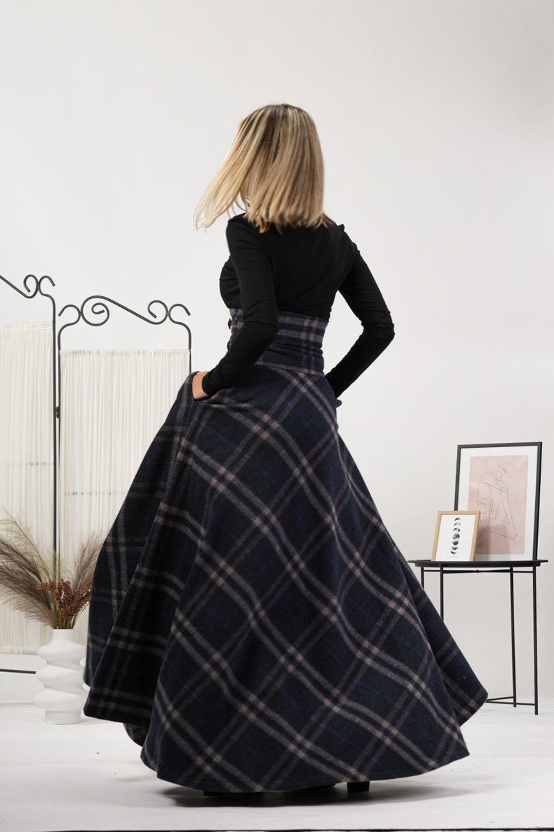 May include: A navy blue plaid skirt with a high waist and a full, flowing design. The skirt has a classic plaid pattern in navy, grey, and tan. The model is wearing a black long-sleeved top. The skirt's length is visible.