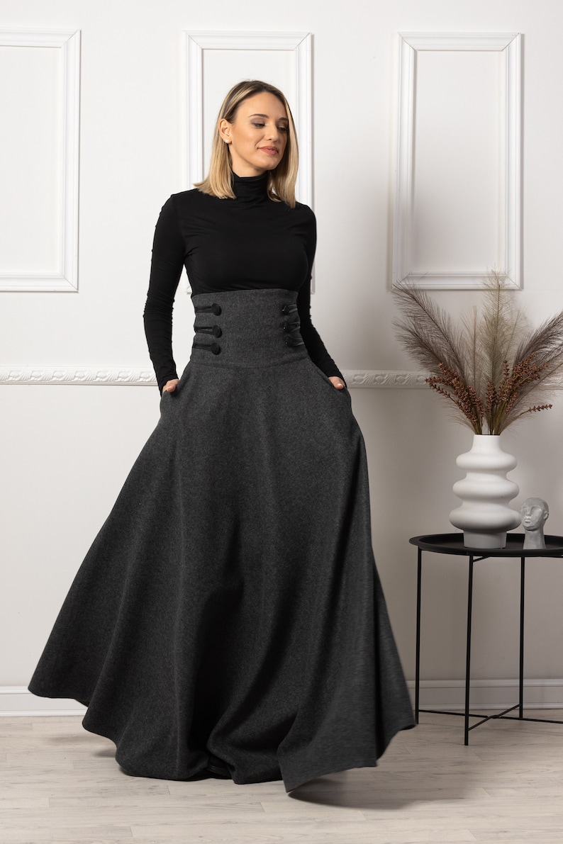 May include: A charcoal grey, high-waisted maxi skirt with large black buttons and pockets. The skirt is paired with a black turtleneck. The skirt's design is a-line, and the fabric appears to be a wool blend.