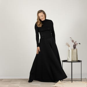 May include: A black long-sleeved dress with a high neckline and a fitted bodice. The bodice has decorative buttons. The skirt is a full, flowing style. The dress is a solid black colour.