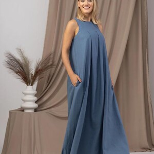 K&ouml;nnte beinhalten: Ein &auml;rmelloses, bodenlanges blaues Kleid mit Falten am Oberteil und Taschen. Das Kleid ist aus einem weichen Stoff gefertigt und hat eine lockere Passform. Die Person l&auml;chelt vor einem neutralen Hintergrund.