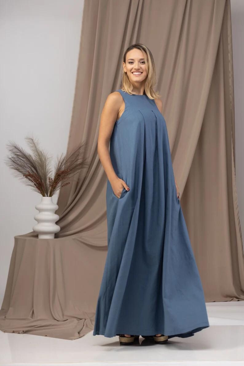 K&ouml;nnte beinhalten: Ein &auml;rmelloses, bodenlanges blaues Kleid mit Falten am Oberteil und Taschen. Das Kleid ist aus einem weichen Stoff gefertigt und hat eine lockere Passform. Die Person l&auml;chelt vor einem neutralen Hintergrund.