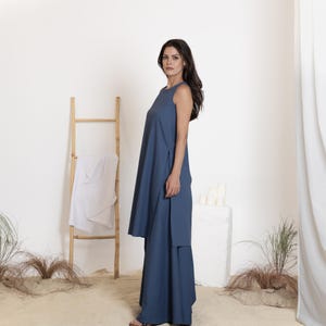 May include: A sleeveless, blue dress with a long, flowing silhouette. The dress features a high neckline and side slits. A wooden ladder with a white cloth draped over it is in the background. The setting is a sandy area with sparse greenery.