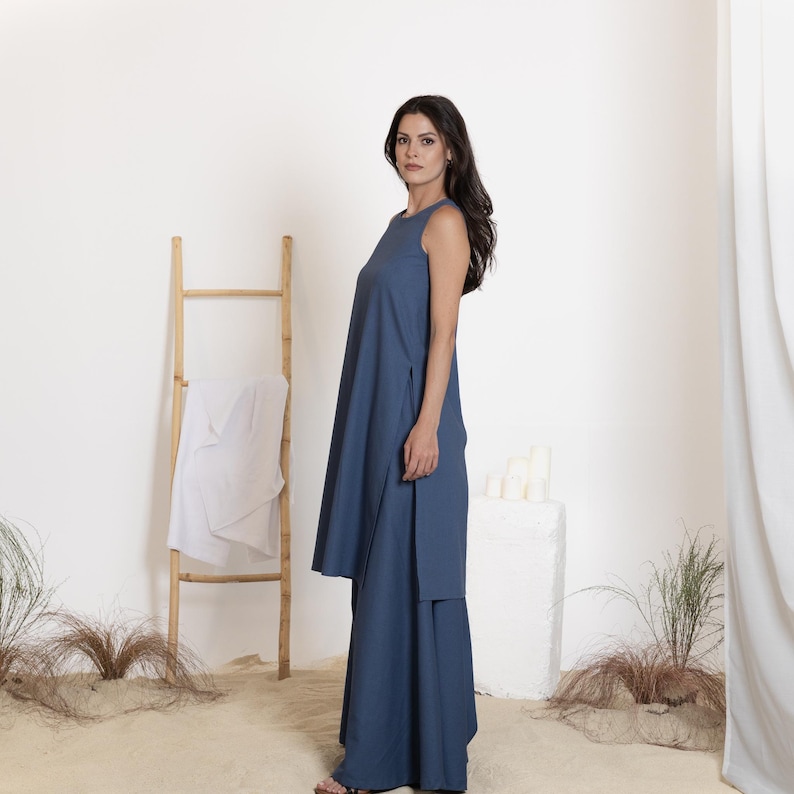 May include: A sleeveless, blue dress with a long, flowing silhouette. The dress features a high neckline and side slits. A wooden ladder with a white cloth draped over it is in the background. The setting is a sandy area with sparse greenery.