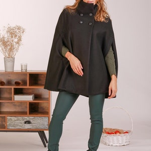 May include: A black wool-blend cape coat featuring a high collar, double-breasted button closure with four round black buttons, and arm slits. The garment is worn over a dark olive green long-sleeved ribbed sweater and dark green fitted trousers.