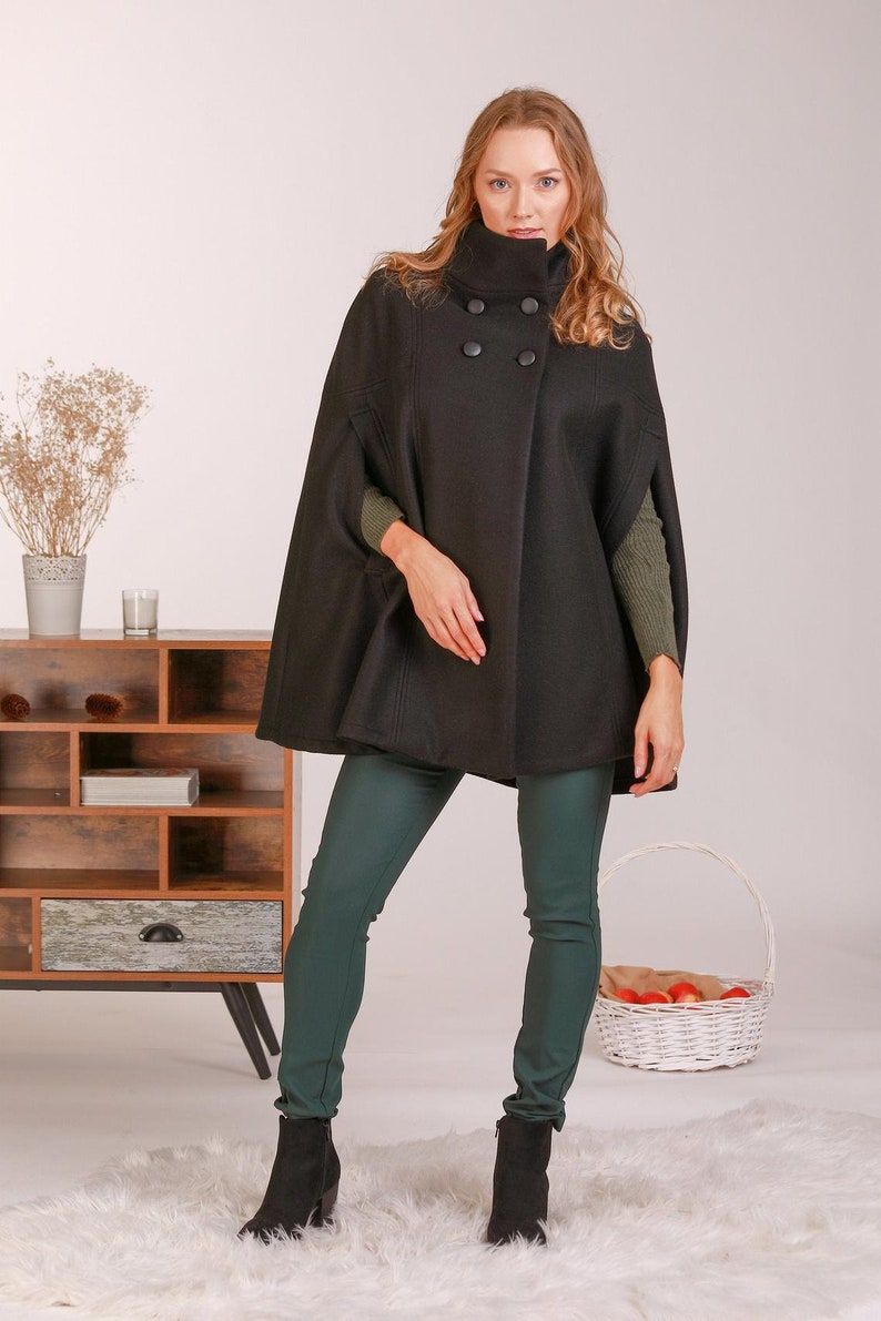 May include: A black wool-blend cape coat featuring a high collar, double-breasted button closure with four round black buttons, and arm slits. The garment is worn over a dark olive green long-sleeved ribbed sweater and dark green fitted trousers.