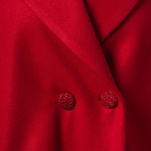 May include: Close-up of a bright red blazer with two matching textured buttons. The blazer's fabric has a smooth, matte finish. The collar is folded over, revealing a glimpse of a black underlayer.