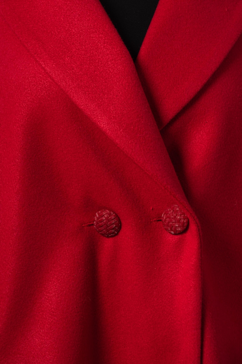 May include: Close-up of a bright red blazer with two matching textured buttons. The blazer's fabric has a smooth, matte finish. The collar is folded over, revealing a glimpse of a black underlayer.