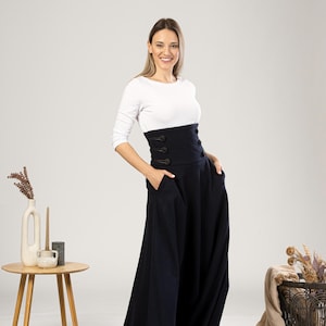 May include: A woman in a white long-sleeved top and a navy blue high-waisted skirt. The skirt has a wide waistband with three buttons and pockets. The outfit is set in a studio with neutral colours.
