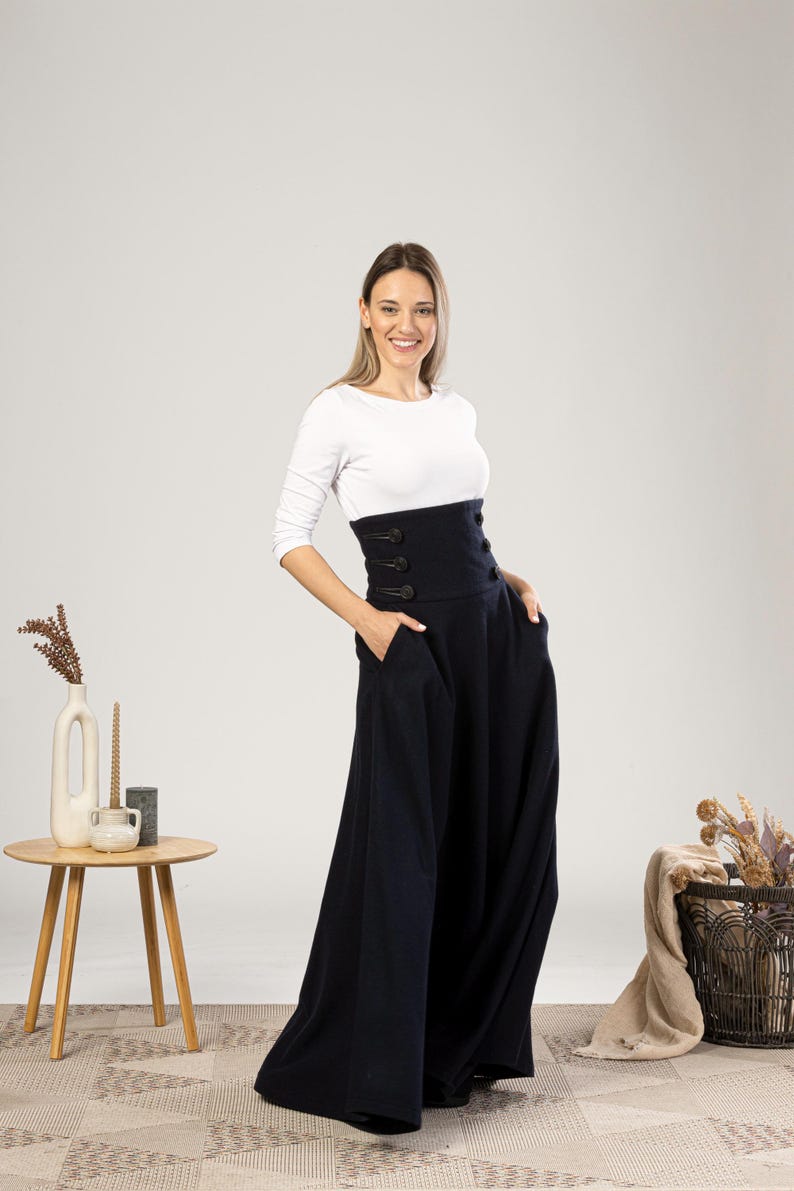 May include: A woman in a white long-sleeved top and a navy blue high-waisted skirt. The skirt has a wide waistband with three buttons and pockets. The outfit is set in a studio with neutral colours.