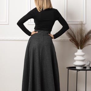 May include: A long, gray skirt with a high waist and a flared silhouette. The skirt is paired with a black long-sleeved top. The model is standing with their hands on their hips. The background includes a white wall and a small black side table.