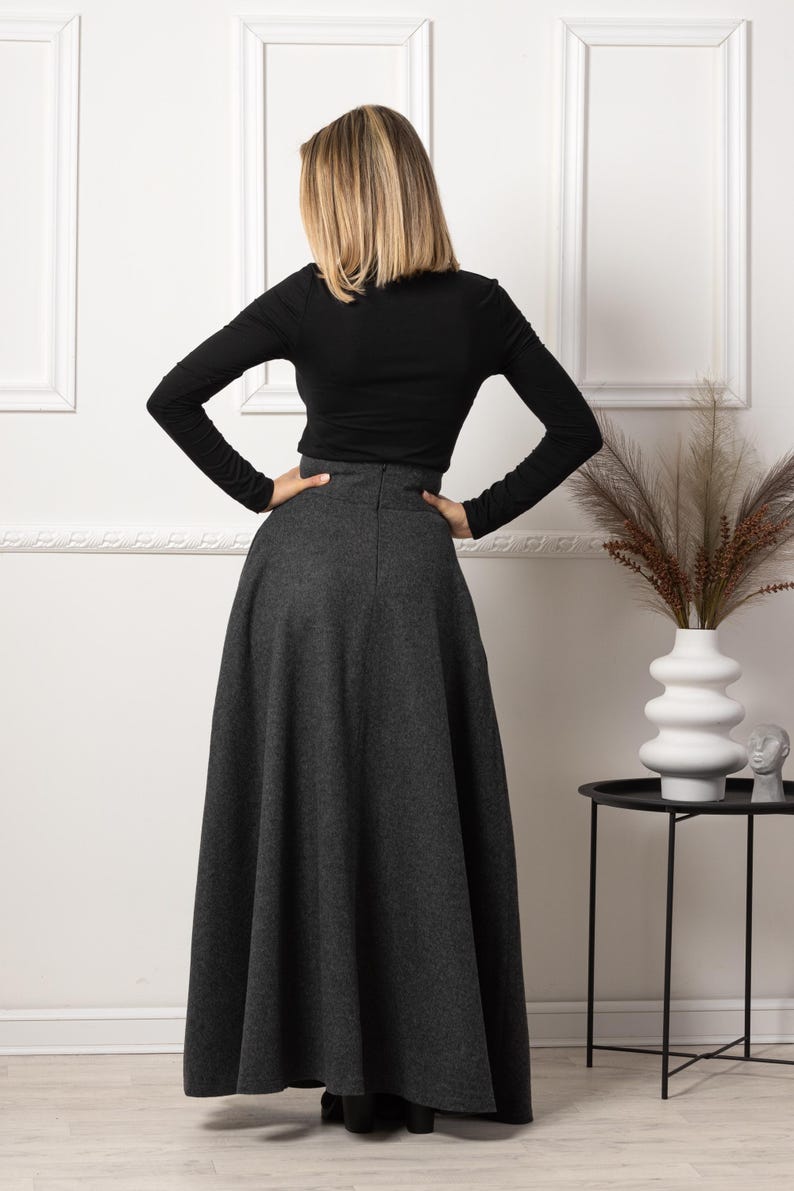 May include: A long, gray skirt with a high waist and a flared silhouette. The skirt is paired with a black long-sleeved top. The model is standing with their hands on their hips. The background includes a white wall and a small black side table.