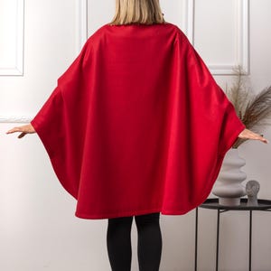 May include: A vibrant red cape-style garment with wide sleeves. The cape is worn over black leggings and black ankle boots. The person is standing in front of a white wall with decorative frames.