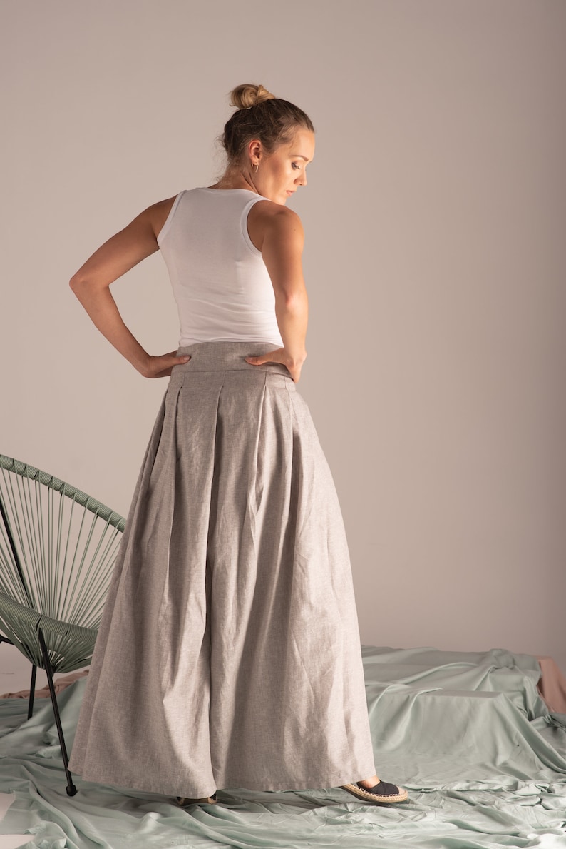 May include: A light grey, pleated maxi skirt and a white vest top. The skirt has a high waist and a full, flowing silhouette. The model is wearing black flats. The background is a neutral colour.