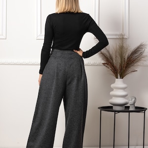 May include: A person wearing a black long-sleeve shirt and gray wide-leg pants. The pants have a high waist and a pleated front. The outfit is paired with a black side table and a white vase with decorative plants.
