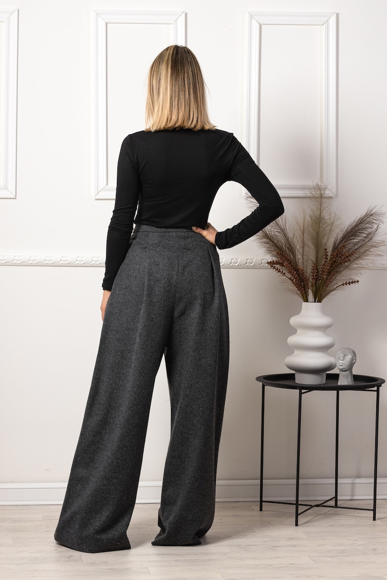 May include: A person wearing a black long-sleeve shirt and gray wide-leg pants. The pants have a high waist and a pleated front. The outfit is paired with a black side table and a white vase with decorative plants.