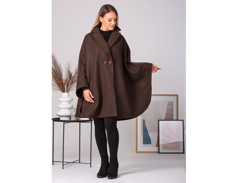 May include: A brown cape coat with a wide collar and two large buttons. The coat has a flowing, rounded shape and is made of a thick, warm material. The model is wearing black leggings and boots.