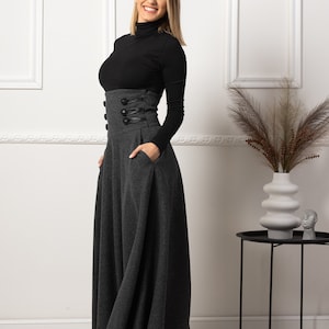 May include: A long, grey skirt with a high waist and decorative buttons. The skirt has a full, flowing shape and pockets. The model is wearing a black polo neck. The skirt is a stylish piece of clothing.