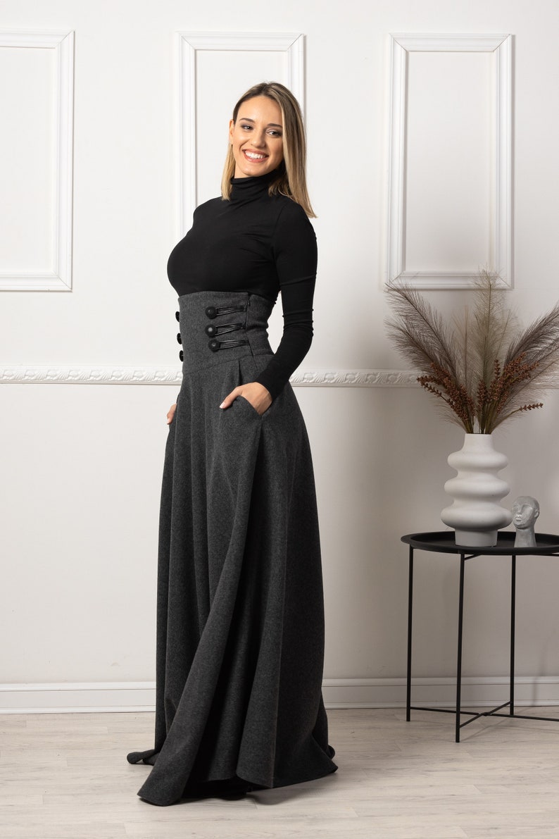 May include: A long, grey skirt with a high waist and decorative buttons. The skirt has a full, flowing shape and pockets. The model is wearing a black polo neck. The skirt is a stylish piece of clothing.
