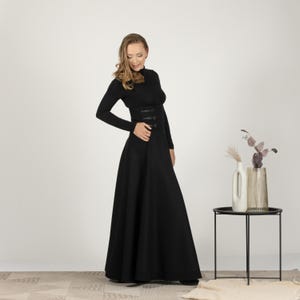 May include: A full-length black dress with a high neck and long sleeves. The dress has a fitted bodice with decorative buttons and a flared skirt. The woman is wearing the dress. The dress is a classic and elegant style.
