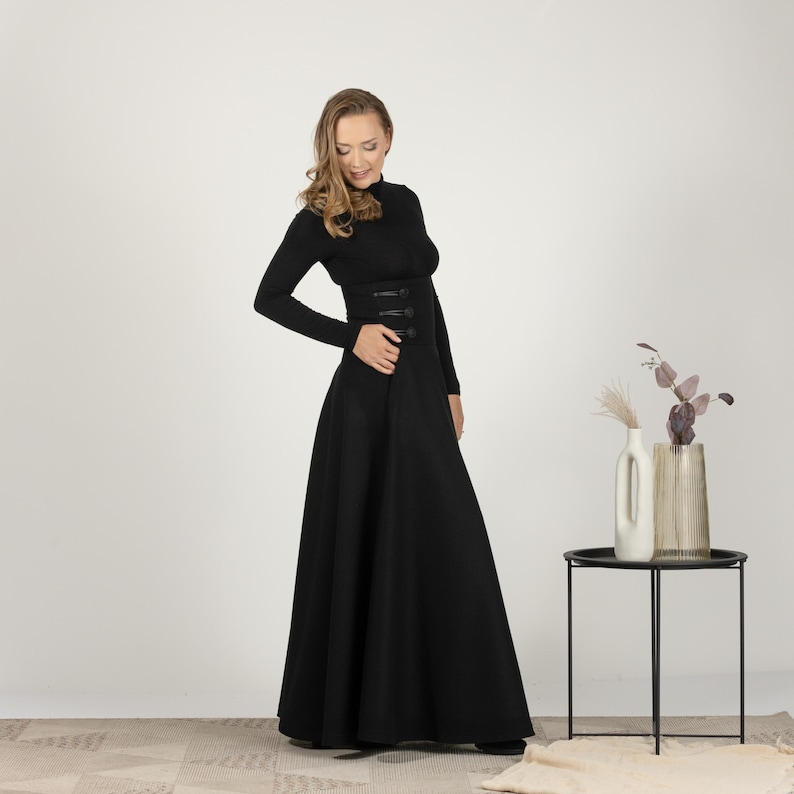 May include: A full-length black dress with a high neck and long sleeves. The dress has a fitted bodice with decorative buttons and a flared skirt. The woman is wearing the dress. The dress is a classic and elegant style.