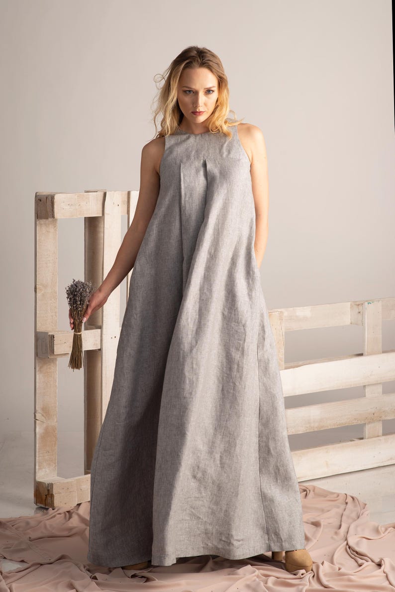K&ouml;nnte beinhalten: Ein hellgraues, &auml;rmelloses Maxikleid mit hohem Ausschnitt. Das Kleid ist aus einem weichen, flie&szlig;enden Stoff gefertigt und hat eine lockere Passform. Das Model h&auml;lt ein B&uuml;ndel getrockneten Lavendels. Das Kleid eignet sich f&uuml;r l&auml;ssige oder formelle Anl&auml;sse.