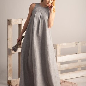 K&ouml;nnte beinhalten: Ein hellgraues, &auml;rmelloses Maxikleid mit einem plissierten Oberteil. Das Kleid ist aus einem leinenartigen Stoff gefertigt und f&auml;llt bis zum Boden. Eine Frau h&auml;lt ein B&uuml;ndel getrockneten Lavendels. Der Hintergrund umfasst eine Holzpalette.