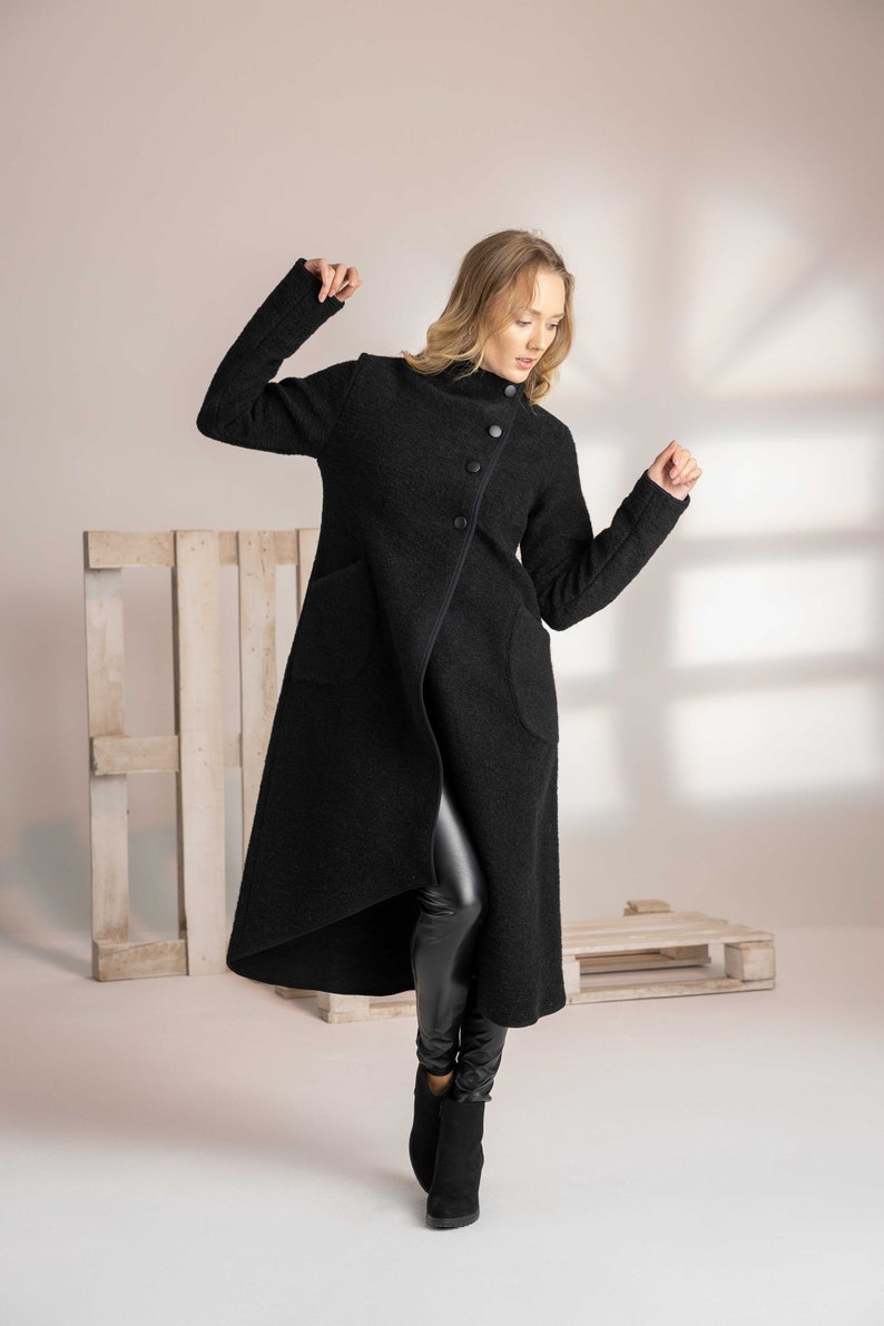 May include: A long, black coat with a unique asymmetrical design. The coat features a high collar, large buttons, and two front pockets. The model is wearing black leather leggings and black boots. The coat is made of a textured fabric.