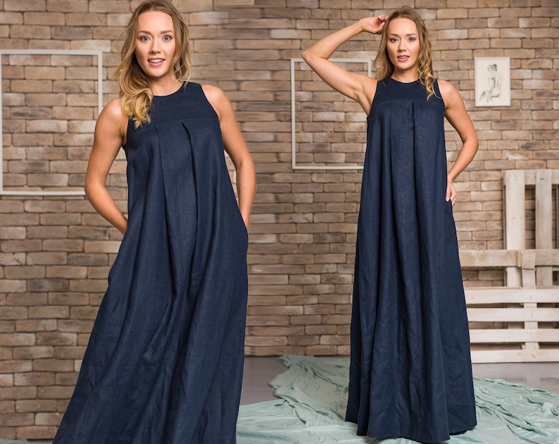 K&ouml;nnte beinhalten: Ein &auml;rmelloses, marineblaues Maxikleid mit plissiertem Oberteil und Taschen. Das Kleid hat eine lockere Passform und einen bodenlangen Saum. Das Model tr&auml;gt das Kleid vor einer Backsteinmauer.