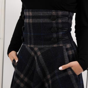 May include: A navy, grey, and brown plaid skirt with a high waist and flared shape. The skirt has a wide waistband with three decorative buttons and pockets. The model is wearing a black long-sleeved top.