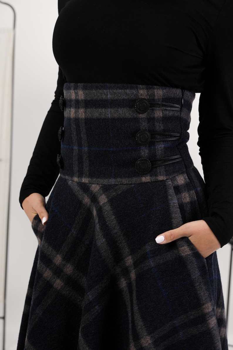 May include: A navy, grey, and brown plaid skirt with a high waist and flared shape. The skirt has a wide waistband with three decorative buttons and pockets. The model is wearing a black long-sleeved top.