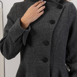 May include: A dark grey wool-blend coat featuring a high collar, long sleeves with folded cuffs, and a flared peplum waist. The coat is fastened with a row of four circular black textured buttons arranged in an asymmetrical diagonal line.