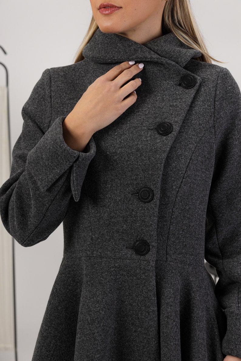 May include: A dark grey wool-blend coat featuring a high collar, long sleeves with folded cuffs, and a flared peplum waist. The coat is fastened with a row of four circular black textured buttons arranged in an asymmetrical diagonal line.