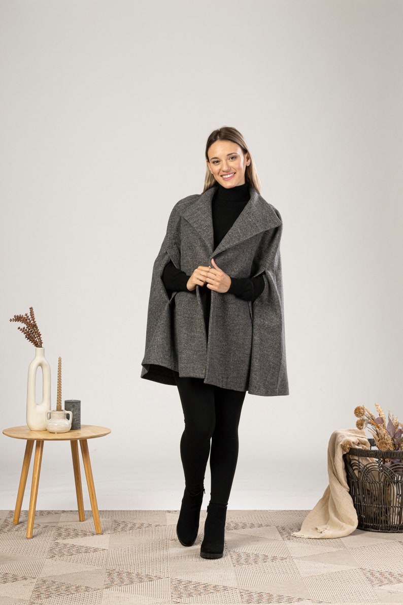 May include: A charcoal gray wool-blend cape featuring a wide collar and open sides, worn over a black long-sleeved turtleneck sweater and black leggings. The garment has a textured finish and falls to the mid-thigh, providing a structured silhouette.