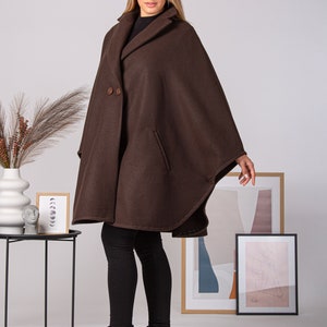 May include: A brown cape coat with a classic design. The coat features a wide collar, two large buttons, and a pocket on the front. The model is wearing black pants and black boots. The coat is made of a soft, warm material.