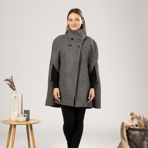 May include: A grey wool cape with a high collar and double-breasted button closure. The cape is knee-length and has a textured appearance. The model is wearing black leggings and boots. A small wooden table and decorative items are in the background.