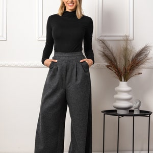 May include: A woman wearing a black turtleneck top and wide-leg gray pants. The pants have a high waist and pockets. The outfit is paired with black shoes. The background is a white wall with decorative frames.