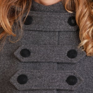 May include: A close-up of a gray wool coat with a high collar and black buttons. The coat has a double-breasted closure and a textured fabric.