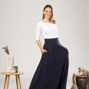 May include: A woman wearing a navy blue maxi skirt with pockets and a white long-sleeve top. The skirt is floor-length and has a high waist. The woman is smiling and has her hand in one of the skirt's pockets.