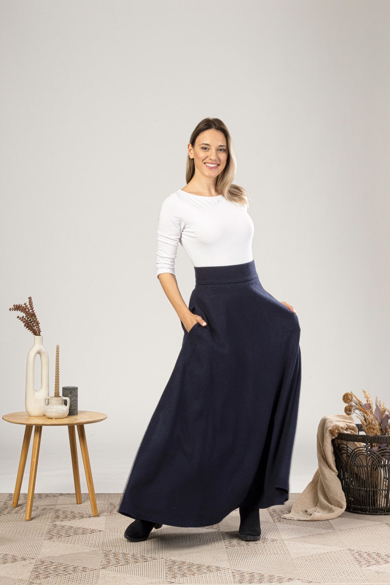 May include: A woman wearing a navy blue maxi skirt with pockets and a white long-sleeve top. The skirt is floor-length and has a high waist. The woman is smiling and has her hand in one of the skirt's pockets.