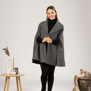 May include: A grey cape with a wide collar. The cape is worn over a black turtleneck and black leggings. The model is wearing black ankle boots. A small wooden table with a vase and candles is to the left.