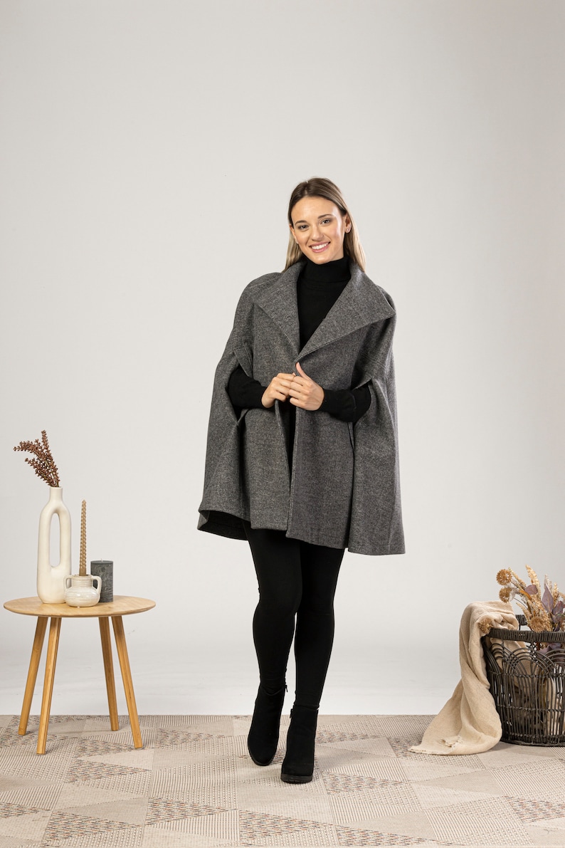 May include: A grey cape with a wide collar. The cape is worn over a black turtleneck and black leggings. The model is wearing black ankle boots. A small wooden table with a vase and candles is to the left.