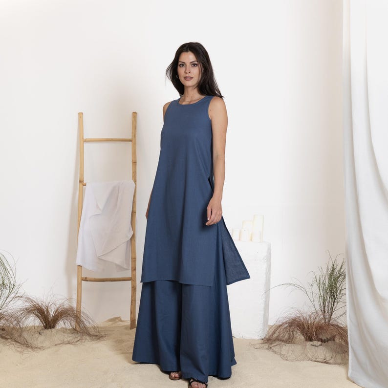 May include: A sleeveless, denim blue outfit consisting of a long tunic over wide-leg pants. The tunic has a high neckline and side slits. The pants are floor-length. The outfit is worn with black sandals. A wooden ladder and white fabric are in the background.
