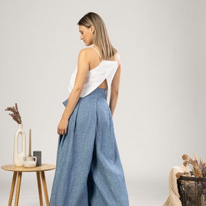 May include: A woman wearing a white top with spaghetti straps and a back cutout, paired with wide-leg, high-waisted blue trousers. The outfit is made of linen-like fabric. A small wooden table with decor is in the background.