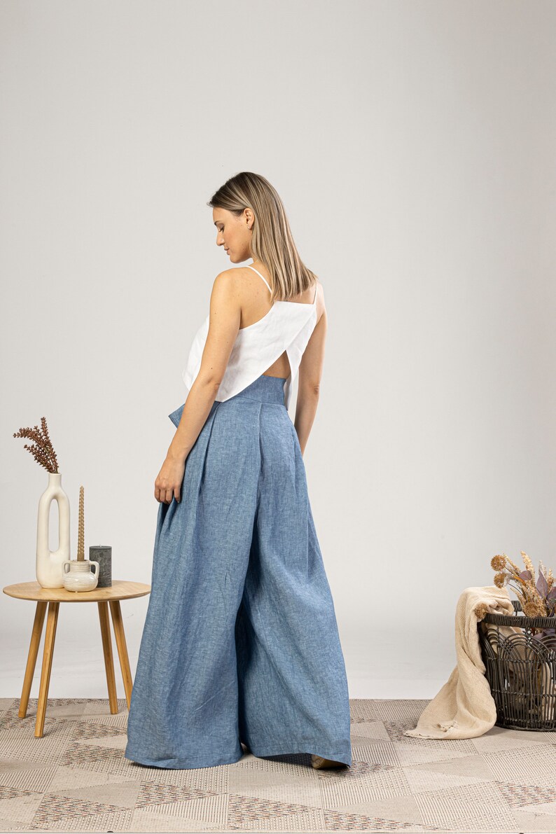 May include: A woman wearing a white top with spaghetti straps and a back cutout, paired with wide-leg, high-waisted blue trousers. The outfit is made of linen-like fabric. A small wooden table with decor is in the background.