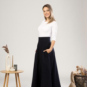 May include: A woman wearing a white long-sleeve shirt and a navy blue, high-waisted, flared skirt with pockets. The skirt falls to the ankles. A small wooden table with a vase and candles is to the left.
