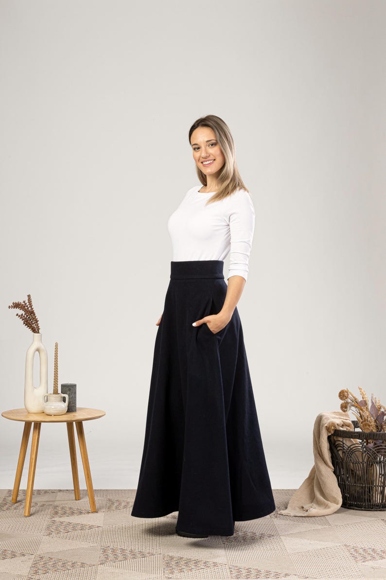 May include: A woman wearing a white long-sleeve shirt and a navy blue, high-waisted, flared skirt with pockets. The skirt falls to the ankles. A small wooden table with a vase and candles is to the left.